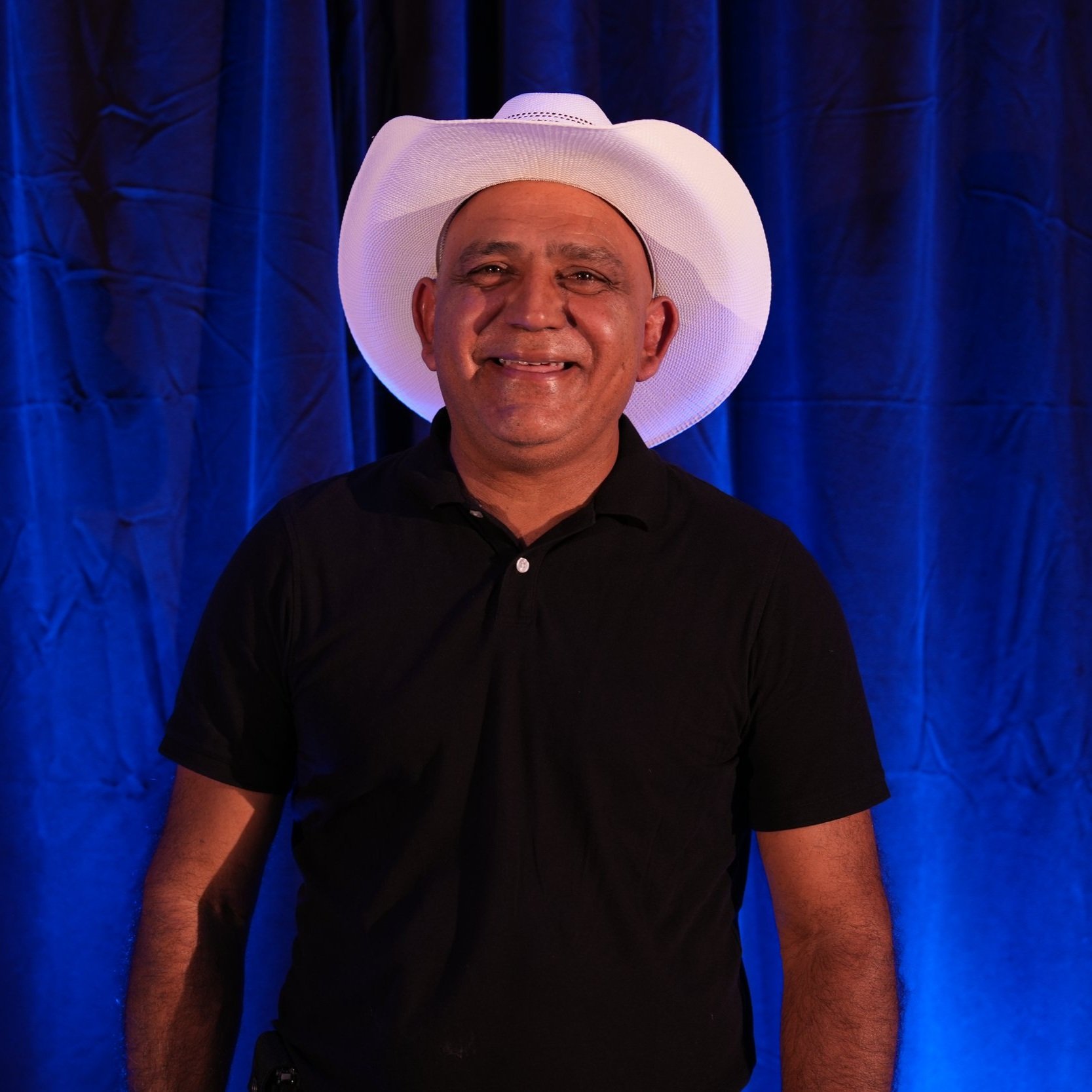WBSAV team member smiling in front of blue curtain backdrop, wearing a white cowboy hat and black polo shirt, capturing personality and pride in a fun, professional setting.